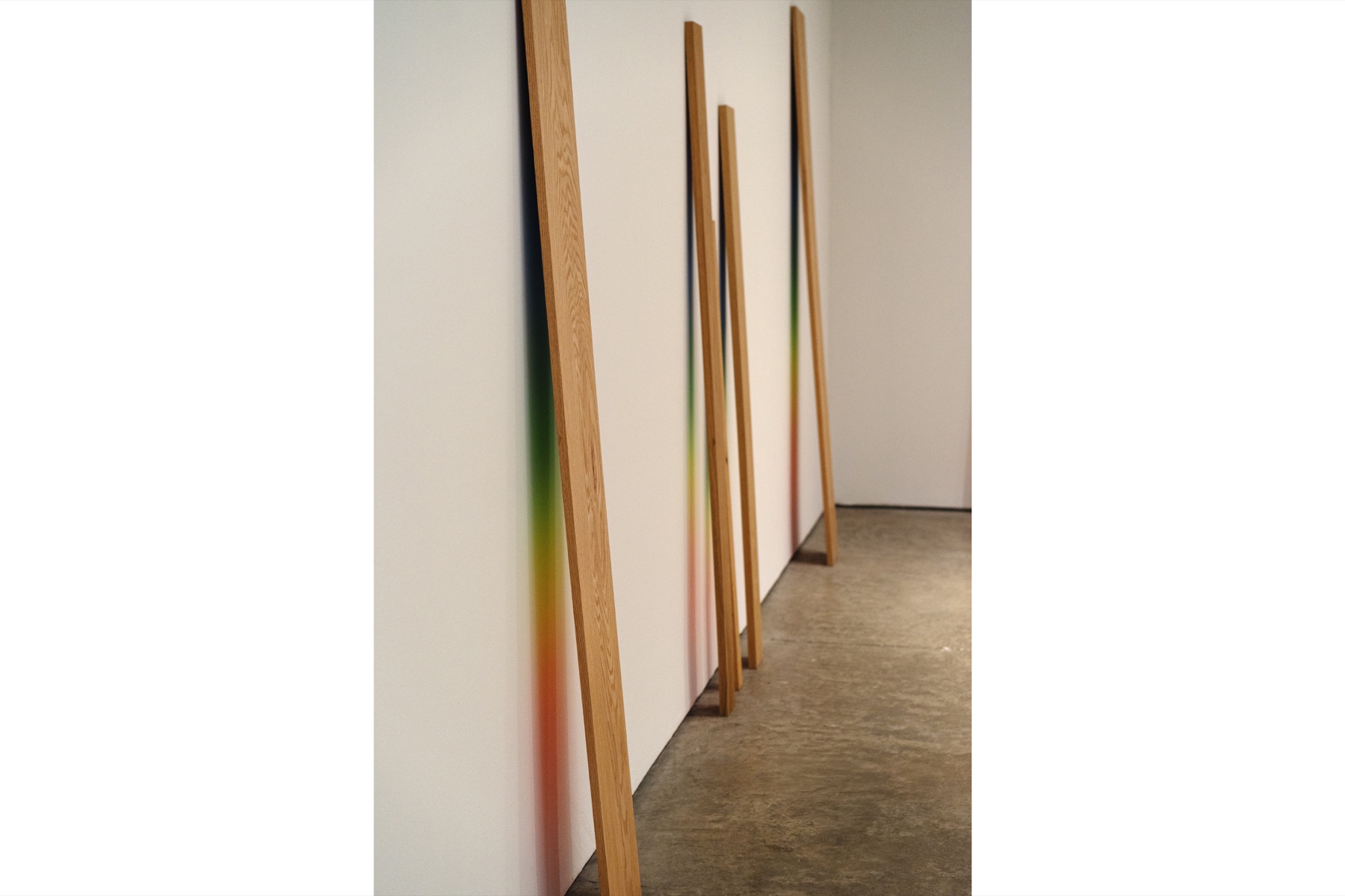 Slender wooden forms cast narrow bands of rainbow light down a white gallery wall.