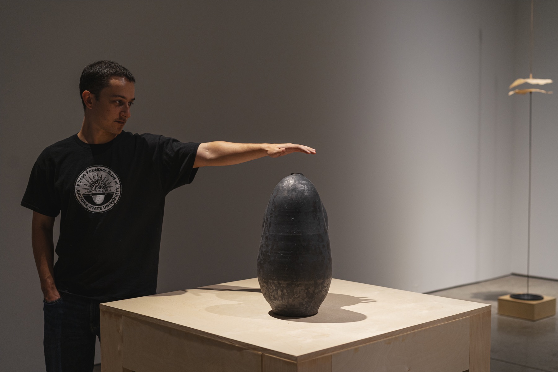 A visitor reaches toward a dark sculptural vessel in the gallery.