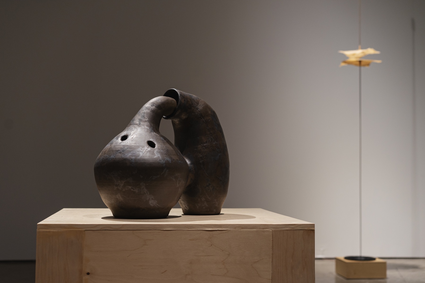 Two joined dark sculptural vessels sit on a pale wood plinth.