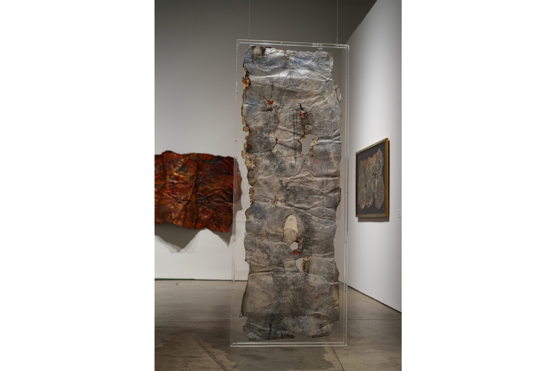 A weathered translucent work hangs vertically inside a clear frame.