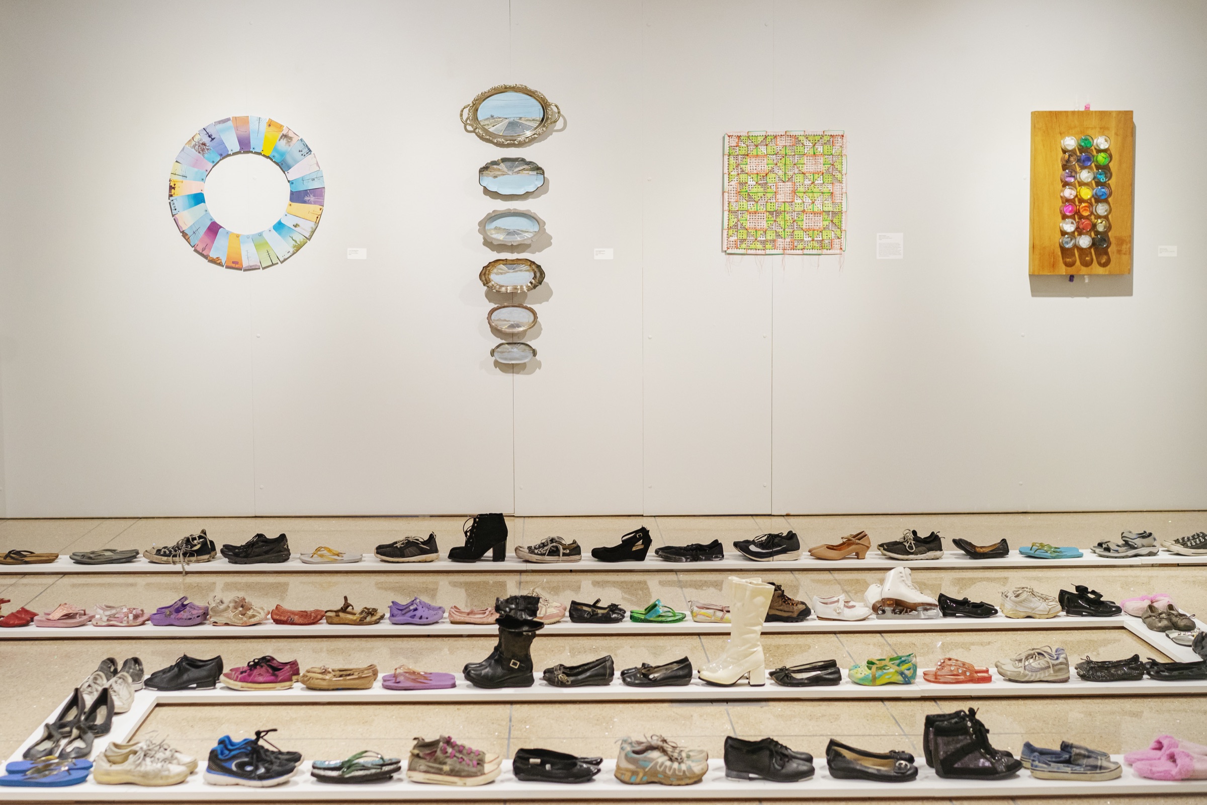 Rows of shoes trace a path across the Vision Gallery floor beneath artworks by Heidi Dauphin and Laura Spaulding Best.