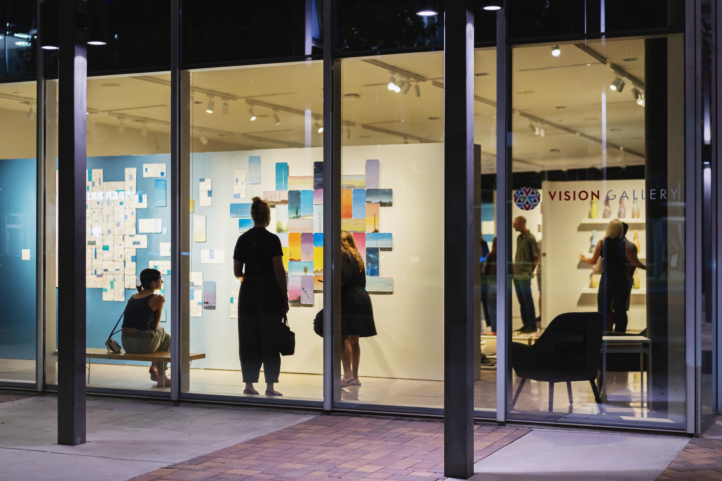 Vision Gallery glows at night as visitors view The Collectionists through its glass storefront.